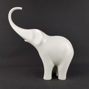 May include: A glossy white ceramic elephant figurine with its trunk raised. The elephant has a smooth, rounded body and four sturdy legs. The figurine is a decorative item, suitable for home decor.