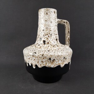 May include: A ceramic vase with a handle, featuring a speckled white and brown upper section and a solid black base. The vase has a bulbous body and a cylindrical neck, with a curved handle on the side. The design is a mix of textures and colors.