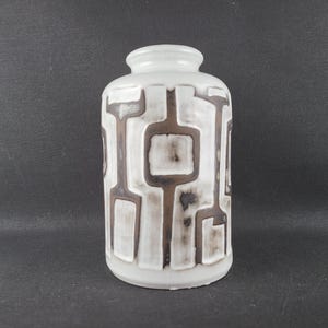 May include: A cylindrical white ceramic vase featuring a raised, dark brown geometric pattern of interconnected lines and rectangular shapes. The vase has a narrow neck and a smooth, glossy finish, with the dark brown design showing subtle shading.