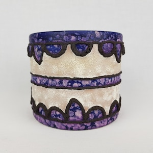May include: A white ceramic planter with a purple and black geometric design. The planter has a textured surface and a rounded shape.