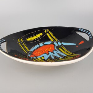 May include: Oval black ceramic serving dish with a white rim and handles. The dish features a colorful abstract design with yellow, orange, and blue shapes. The handles have a striped design.