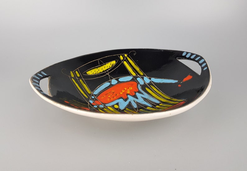 May include: Oval black ceramic serving dish with a white rim and handles. The dish features a colorful abstract design with yellow, orange, and blue shapes. The handles have a striped design.