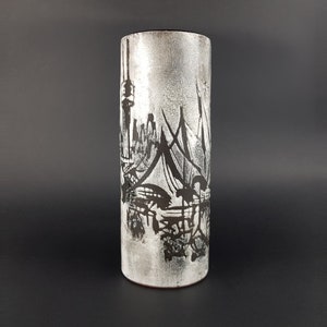 Może przedstawiać: Cylindryczny wazon z teksturowaną, srebrzystą powierzchnią. Wazon ma czarny, abstrakcyjny wzór krajobrazu miejskiego. Projekt zawiera elementy architektoniczne i odbicia, tworząc uderzający kontrast.
