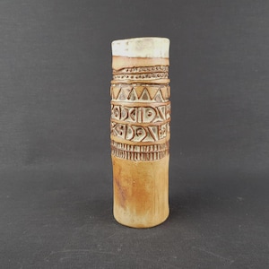 May include: A tall, cylindrical ceramic vase with a textured surface and intricate carved patterns. The vase features a light brown base, transitioning to a beige top. Decorative bands with geometric designs encircle the vase.