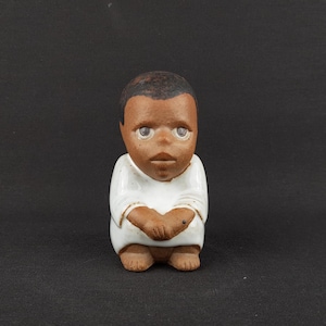 Puede incluir: Pequeña figura de cerámica de una persona con piel oscura, pelo negro corto y ojos grandes. La figura lleva una camisa blanca y tiene brazos y piernas marrones. Las manos están juntas. Sobre un fondo negro.