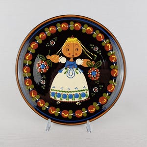 Może przedstawiać: Ozdobny ceramiczny talerz z czarnym tłem i kolorową kwiatową obwódką. Talerz przedstawia ilustrację w stylu sztuki ludowej kobiety w tradycyjnym stroju, trzymającej kwiaty. Talerz jest eksponowany na przezroczystym stojaku.