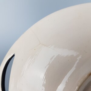 May include: Close-up of a white ceramic plate with a glossy finish. The plate has a crack running across its surface. A black handle is visible on the side of the plate. The plate is set against a light blue background.