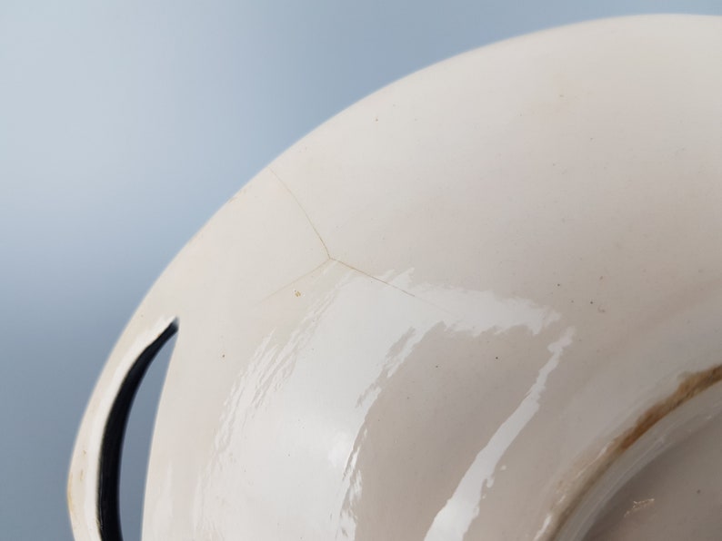 May include: Close-up of a white ceramic plate with a glossy finish. The plate has a crack running across its surface. A black handle is visible on the side of the plate. The plate is set against a light blue background.