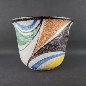 Op de afbeelding: Een keramische plantenpot met een uitlopende rand en een textuurrijke, gespikkelde witte basis. De buitenkant is voorzien van abstracte, gebogen geometrische vormen in blauw, groen, oranje, geel en zwart, wat een veelkleurig patroon vormt.