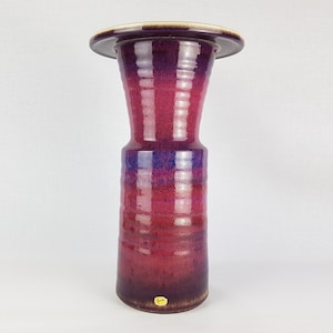 May include: A tall, ceramic vase with a wide, flared opening and a narrow base. The vase is decorated with a gradient of pink, purple, and blue glaze, creating a striped effect. The vase has a glossy finish and a small, gold sticker on the bottom.