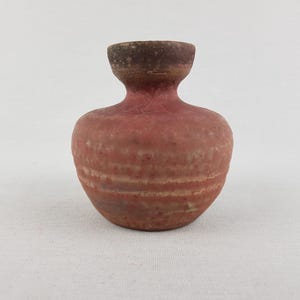 May include: A small, round, terracotta-colored ceramic vase with a narrow neck and a flared rim. The surface features a textured, matte finish with subtle horizontal bands of lighter and darker reddish-brown tones across the bulbous body.