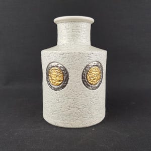 Puede incluir: Un jarrón de cerámica texturizada de color blanco roto, con cuerpo cilíndrico y cuello ligeramente acampanado. Dos medallones ovalados dorados con bordes plateados están centrados en el frente. El jarrón tiene un aspecto moteado.