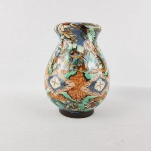 Może przedstawiać: Ozdobny ceramiczny wazon o szerokim korpusie i wąskiej szyi. Wazon ma kolorowy wzór mozaiki z odcieniami niebieskiego, pomarańczowego, zielonego i brązowego. Podstawa jest w kolorze ciemnobrązowym.