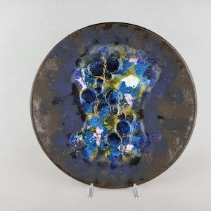 May include: A circular decorative plate featuring a dark brown outer rim and a textured center with abstract patterns in shades of deep blue, bright turquoise, yellow, and white, accented by raised circular bubble-like formations throughout.