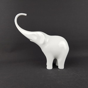 May include: A glossy white elephant figurine with its trunk raised. The ceramic sculpture has a smooth, reflective surface and is set against a dark background. The elephant is a decorative item.