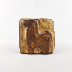 May include: A rectangular ceramic vase with a relief of a stylized animal. The vase is primarily brown with lighter tan accents, and the animal figure is a darker shade of brown. The vase has a rustic, handcrafted appearance.