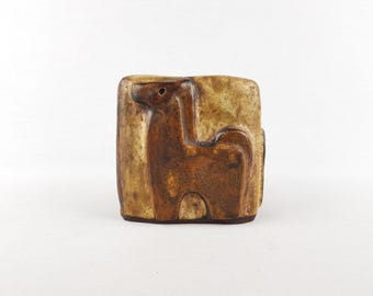 Peut inclure: Un vase rectangulaire en céramique avec un relief d'animal stylisé. Le vase est principalement marron avec des accents beige clair, et la figure de l'animal est d'une teinte plus foncée de marron. Le vase a un aspect rustique et artisanal.