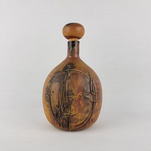 Op de afbeelding: Een bruine keramische fles met een ronde, bruine stop. De fles heeft een getextureerd oppervlak en een ontwerp van een schip met een zeil.