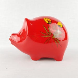 Könnte beinhalten: Ein glänzendes, rotes Keramik-Sparschwein. Das Schwein hat einen abgerundeten Körper, kleine Beine und eine Schnauze. Es ist mit gelben und goldenen floralen Akzenten verziert. Ein klassischer Artikel zum Sparen.