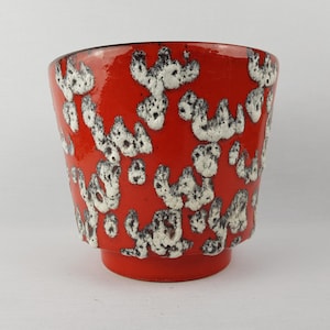 Puede incluir: Una vibrante maceta de cerámica roja con un patrón moteado único en blanco y negro. La maceta tiene una forma ligeramente acampanada y una pequeña base elevada. Una pieza decorativa para plantas de interior.