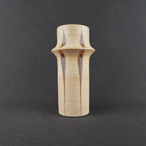 May include: A cylindrical ceramic vase with a textured beige surface and a prominent flared rim. The vase features vertical, elongated teardrop-shaped brown accents that extend downward from the flared section, creating a distinct visual pattern.