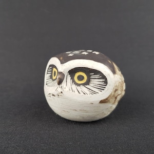 Puede incluir: Pequeña figura de búho pintada a mano. El búho tiene un cuerpo blanco con una cabeza marrón y negra. Los ojos son amarillos con detalles negros. El objeto es decorativo, probablemente para una estantería o escritorio.