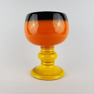 May include: A decorative goblet with a glossy finish. The top is black, transitioning to orange, and the stem and base are yellow. The goblet has a rounded bowl shape and a detailed stem.