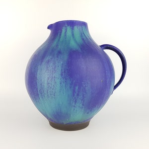 Peut inclure: Un pichet en céramique bleu et vert avec un corps arrondi et une anse. Le pichet a un glaçage marbré avec des nuances de bleu et de vert.