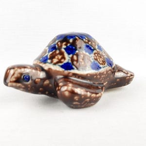 May include: A ceramic turtle ornament. The turtle has a brown body and a shell decorated with blue and brown patterns. The turtle has a blue eye and is a decorative item.