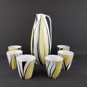May include: A ceramic pitcher and six matching cups. The pitcher is tall and slender, with a black, white, and yellow abstract design. The cups have a similar design. The set is on a dark gray surface.