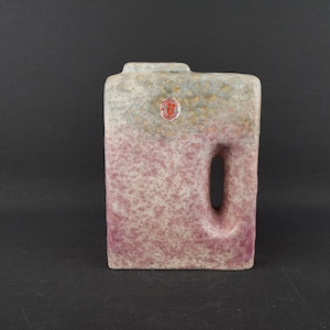 Peut inclure: Un vase en céramique avec une surface texturée et mouchetée dans des tons de gris, rose et brun. Le vase a une forme rectangulaire avec une poignée découpée et un tampon rouge sur le devant.