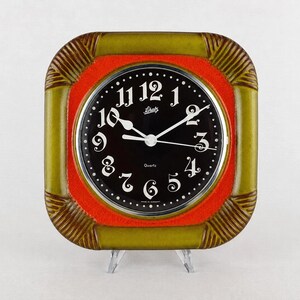 Op de afbeelding: Een vierkante wandklok met een glanzende olijfgroene keramische lijst met geribbelde hoeken en een feloranje binnenrand. De ronde zwarte wijzerplaat toont witte Arabische cijfers, witte wijzers en de tekst Schatz en Made in Germany.