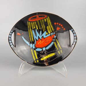 May include: A black ceramic serving dish with a colorful abstract design. The dish features a central design with yellow, blue, and red shapes. It has two handles with blue and white accents. The dish is displayed on a clear stand.