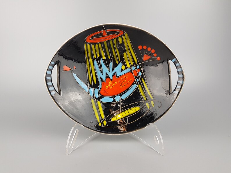 May include: A black ceramic serving dish with a colorful abstract design. The dish features a central design with yellow, blue, and red shapes. It has two handles with blue and white accents. The dish is displayed on a clear stand.