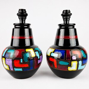 Op de afbeelding: Twee zwarte tafellampen met een geometrisch ontwerp. Elke lamp heeft een zwarte basis en bovenkant met een rode streep. Het onderste deel van elke lamp heeft een kleurrijk mozaïekpatroon met rode, blauwe, gele en paarse vormen.