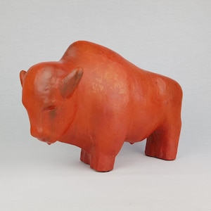 Puede incluir: Una escultura de bisonte de cerámica roja con una superficie texturizada. El bisonte está de pie con la cabeza baja.
