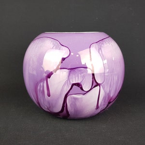 Puede incluir: Un jarrón de vidrio morado con vetas blancas y moradas. El jarrón es redondo y tiene un acabado brillante.