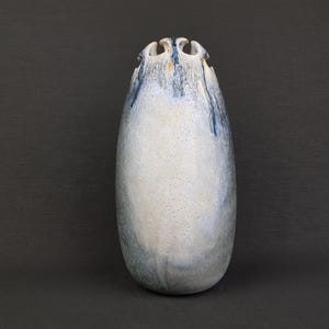 May include: A white ceramic vase with a blue glaze and a unique, scalloped rim. The vase has a speckled texture and a slightly oval shape.
