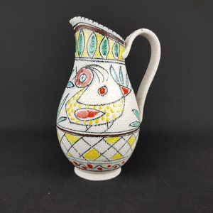 May include: A white ceramic jug with a yellow, red and blue bird design. The jug has a handle and a rounded base.