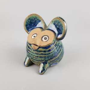 Puede incluir: Una pequeña figura de cerámica con forma de ratón estilizado y orejas grandes y redondeadas. El cuerpo presenta un esmalte brillante a rayas azules y verdes, mientras que la cara es de color beige mate con dos ojos circulares blancos.