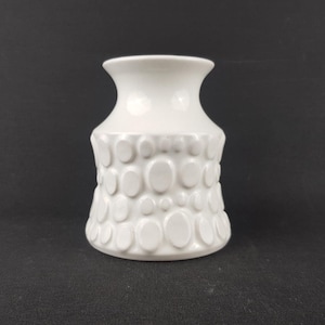 Pode incluir: Um vaso de cerâmica branco com um padrão oval texturizado na parte inferior. O vaso tem um topo alargado e uma base arredondada. O fundo é preto sólido.