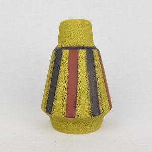 Pode incluir: Um vaso de cerâmica amarela texturizada com uma base alargada e um pescoço cilíndrico. O vaso apresenta listras verticais em preto e castanho, adicionando um padrão geométrico ao design. O vaso é um item decorativo.