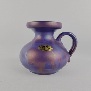 May include: A small, purple ceramic vase with a handle. The vase has a bulbous body and a narrow neck. It features a metallic, iridescent finish with gold accents. The label "Topeko Keramik" is visible on the front.