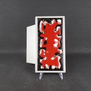 Op de afbeelding: Een rechthoekige keramische wandplaat met een glanzende zwarte achtergrond en een centraal, organisch, verhoogd rood glazuurpatroon met witte randen. Het object is gemonteerd op een doorzichtige acrylstandaard, wat de verticale oriëntatie benadrukt.