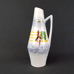 Puede incluir: Una jarra alta de cerámica blanca con asa, decorada con rayas amarillas y formas abstractas coloridas. La jarra tiene un patrón moteado azul y blanco en la mitad inferior. Una pequeña etiqueta está adherida al costado.