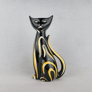 Könnte beinhalten: Eine glänzende schwarze Keramikkatzenfigur mit stilisierten gelben und weißen geschwungenen Linienmustern auf dem Körper. Die Katze ist in sitzender Position mit langem Hals, spitzen Ohren und mandelförmigen Augen dargestellt und hat eine glatte Glasur.