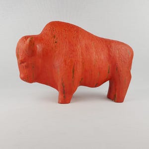 Puede incluir: Escultura artesanal de un bisonte de color naranja. La figura tiene una superficie texturizada con líneas verticales y un ligero brillo. El bisonte está representado de pie, con cuatro patas y una prominente joroba.