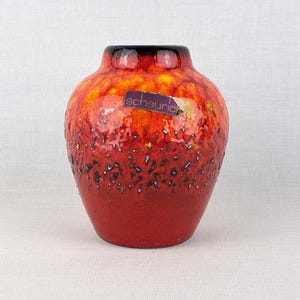 Puede incluir: Un jarrón de cerámica con forma bulbosa, que presenta un degradado vibrante de esmalte rojo, naranja y amarillo. La superficie tiene un acabado texturizado y moteado con acentos oscuros cerca de la base. Una pegatina morada con el texto scheurich está adherida.