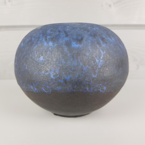 May include: A round, dark gray ceramic vase with a blue glaze. The glaze has a mottled effect, with darker blue areas and lighter blue areas.
