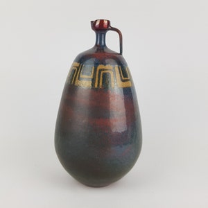 May include: A handcrafted ceramic jug with a unique shape and a handle. The jug features a dark red and blue glaze with gold geometric patterns. The jug's design is both functional and decorative, suitable for display or use.